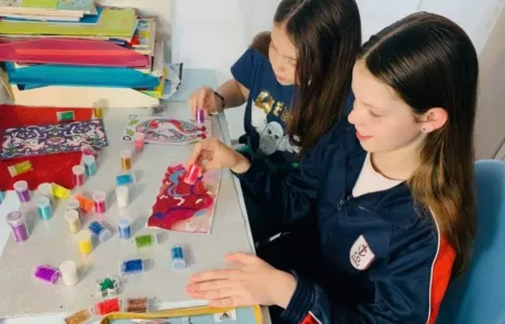 Dos alumnas del COAMI aplican purpurina de colores sobre sus dibujos en clase.