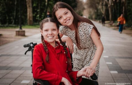 Dos hermanas sonríen en un parque; la mayor, con abrigo rojo, va en silla de ruedas.