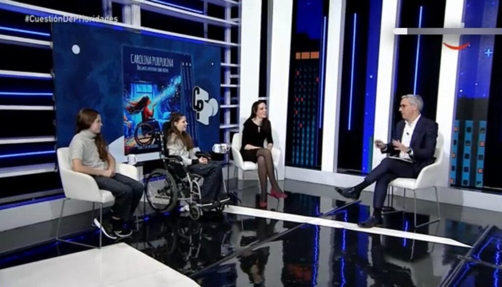 Fernsehstudio: zwei Mädchen, eines im Rollstuhl, und eine Frau unterhalten sich mit dem Moderator vor der Titelseite des Buches.