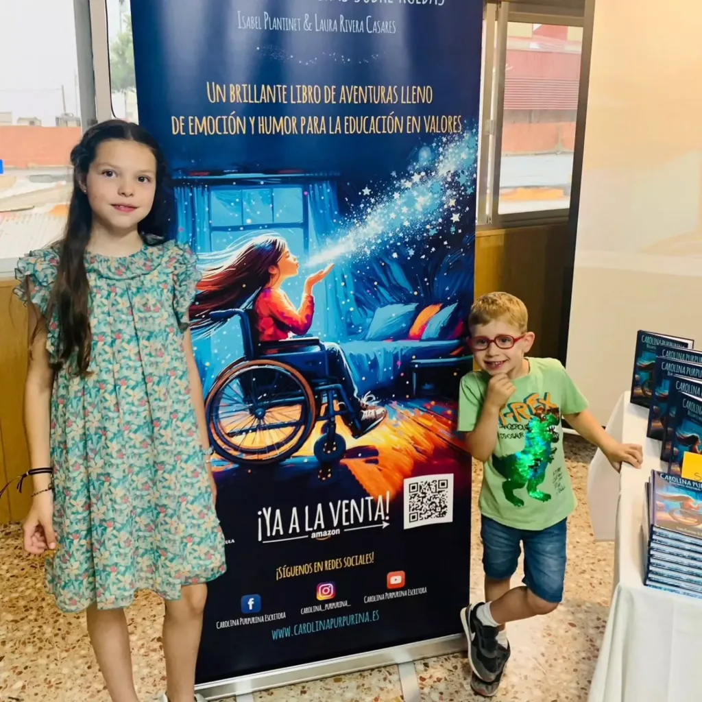 Una niña y un niño con gafas posan junto al roll-up del libro, ante una mesa con ejemplares apilados.