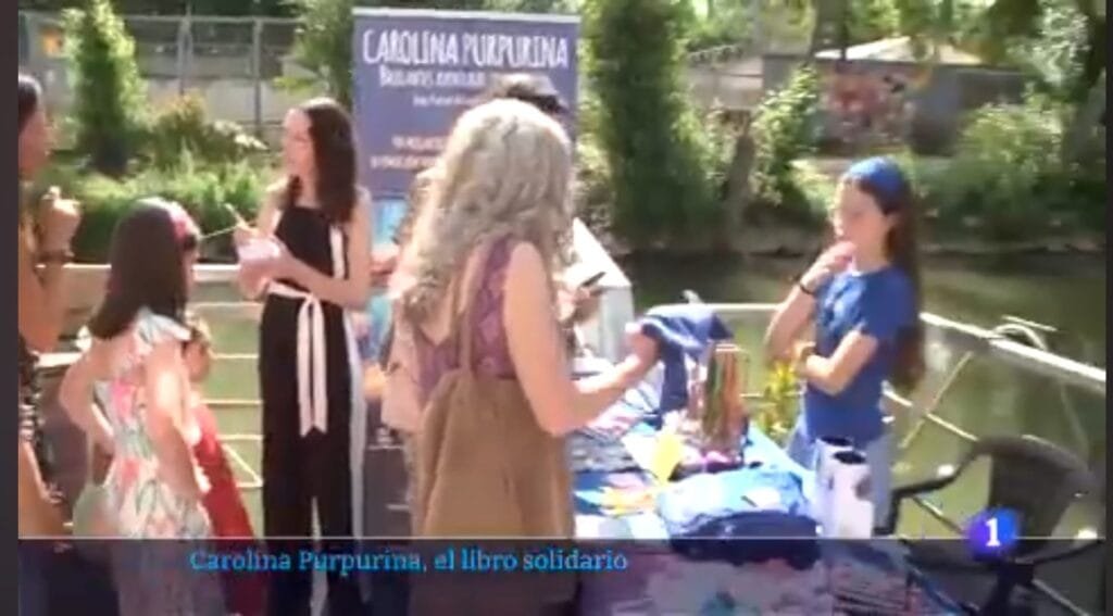 Standbild von TVE mit dem Titel „Carolina Purpurina, el libro solidario“: Besucher vor dem Bücherstand am Fluss.
