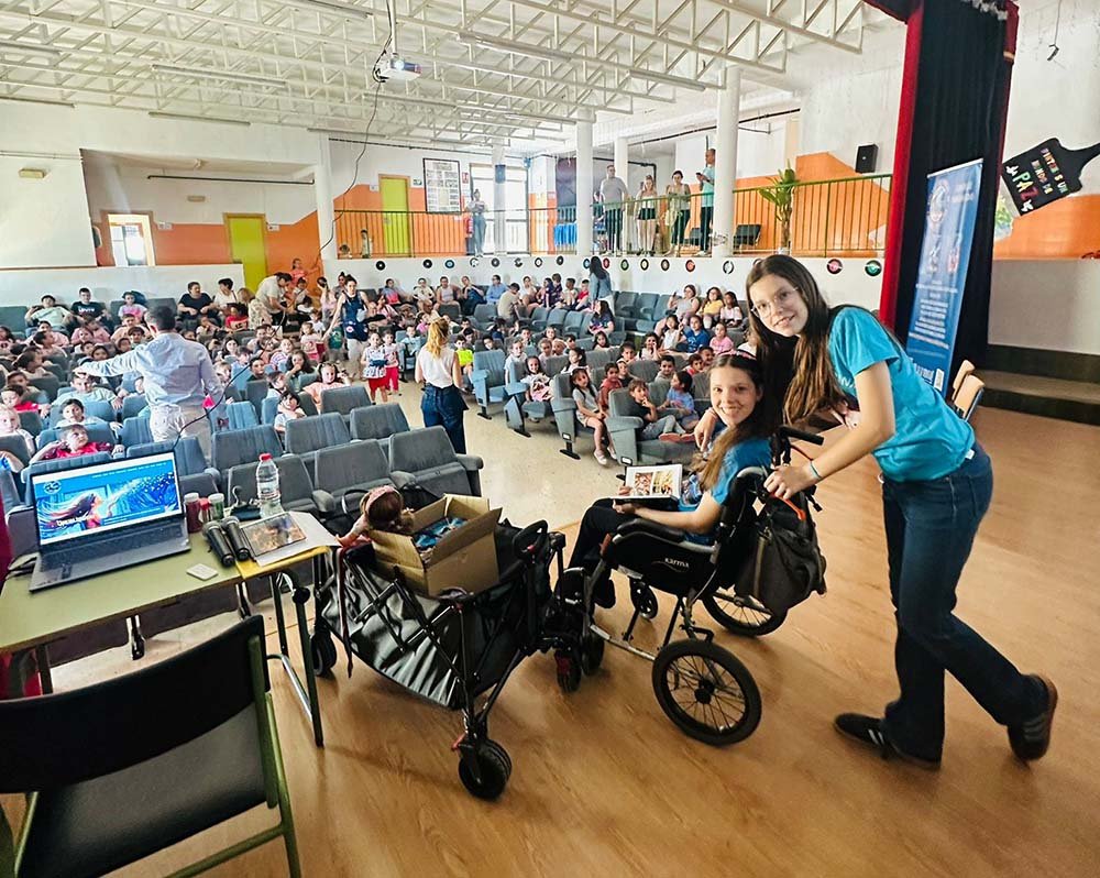 Carolina e Isabel en el salón de actos del colegio, ante el público de alumnos, junto a la caja de libros.