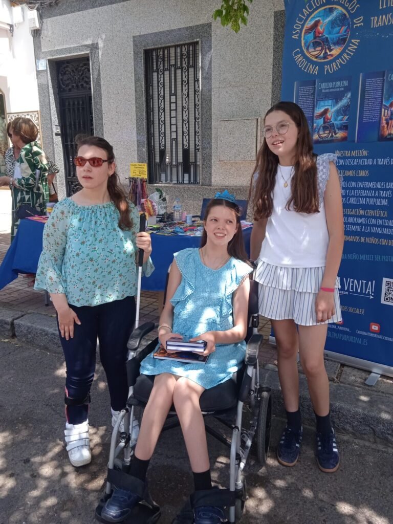 Carolina sostiene los libros en el puesto, acompañada de Isabel y de una lectora con bastón blanco.