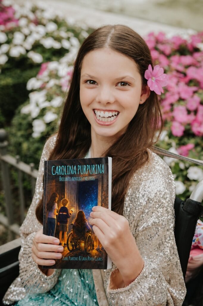 Isabel hält „Carolina Purpurina. Brillantes aventuras y misterios“ vor einem Blumenbeet.