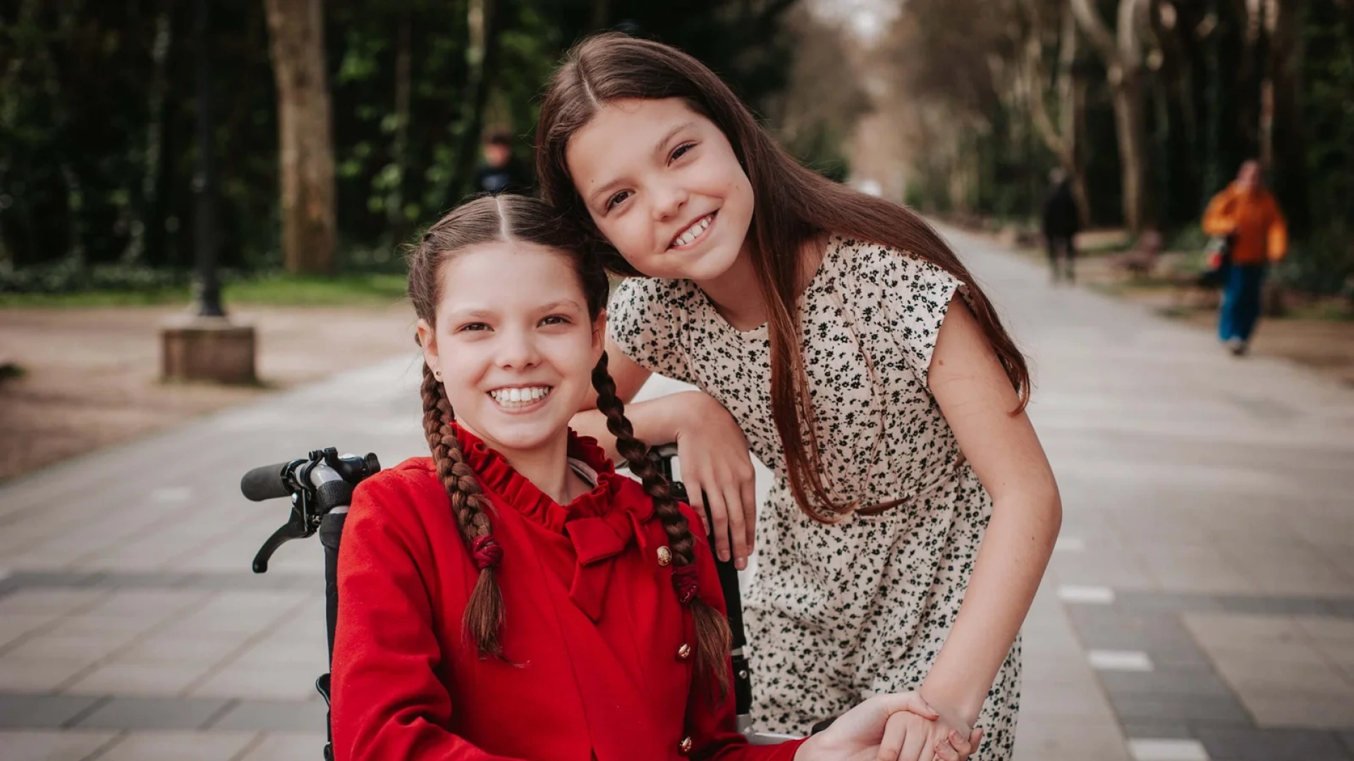 Carolina, con abrigo rojo y trenzas, sonríe en su silla de ruedas junto a otra niña que le da la mano, en un paseo arbolado.