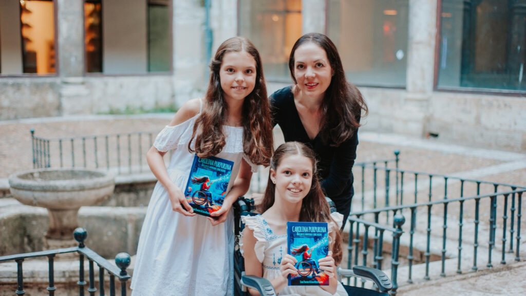Carolina, en su silla de ruedas, y otra niña muestran el libro «Carolina Purpurina» junto a una mujer, en un patio de piedra.