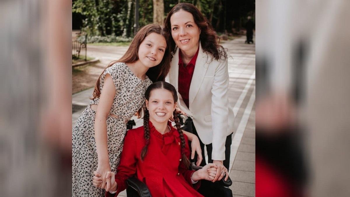 Carolina, con vestido rojo y trenzas, sonríe en su silla de ruedas, junto a una niña de vestido estampado y una mujer de chaqueta blanca.