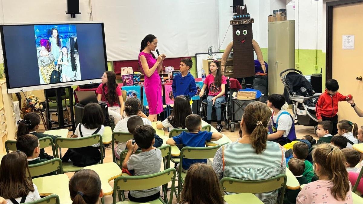 Una mujer habla con micrófono ante los alumnos sentados; a su lado, varios niños y Carolina, en su silla de ruedas, junto a una mesa con libros.