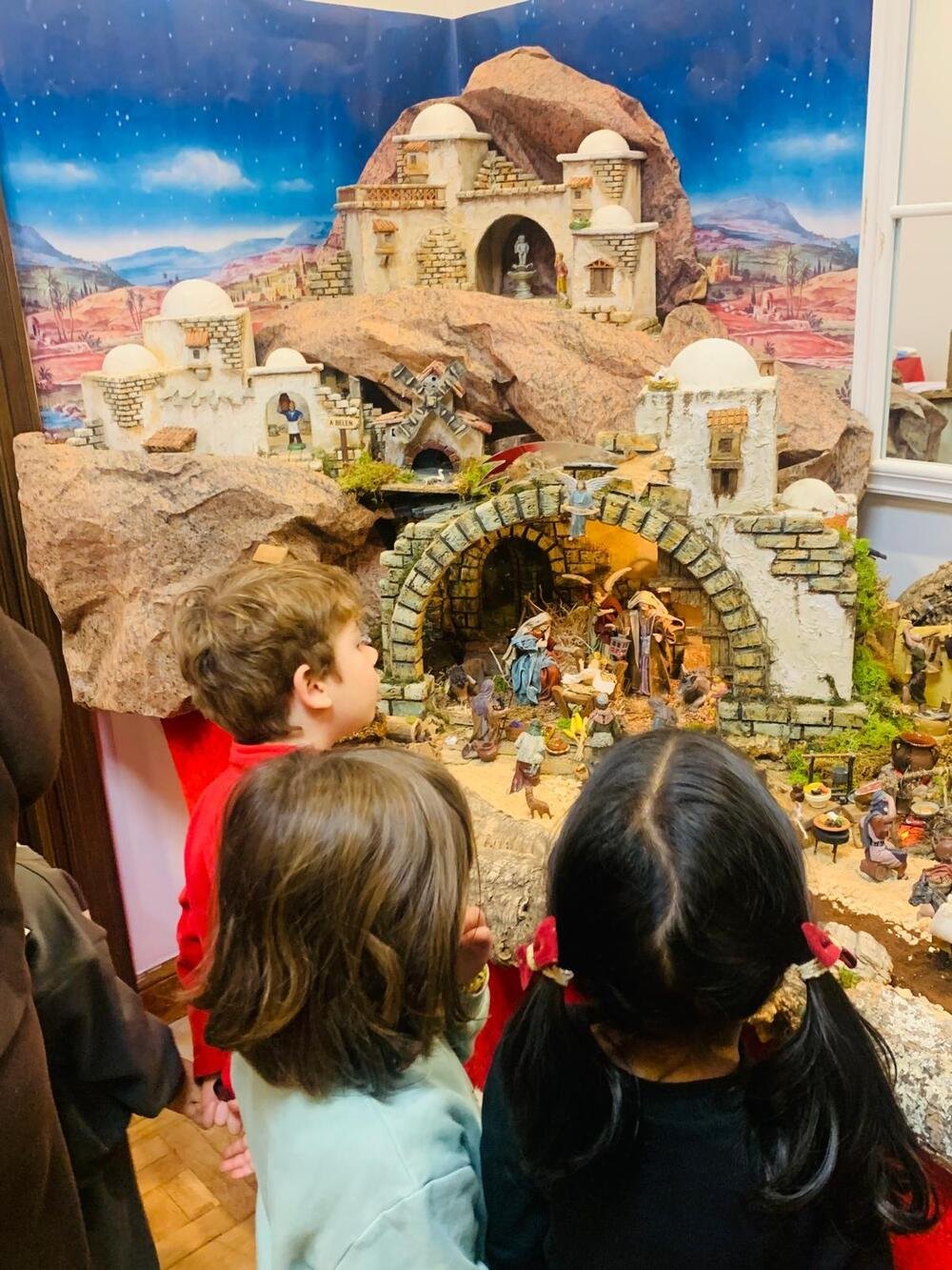 Tres niños, de espaldas, contemplan el belén: casas blancas, un portal de piedra con el nacimiento y un fondo de cielo estrellado.