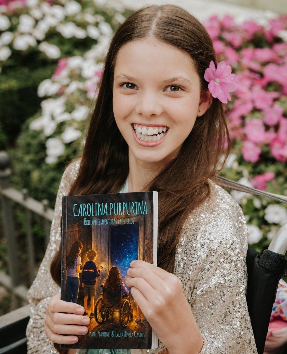 Una niña sonriente, con una flor rosa en el pelo, sostiene el libro «Carolina Purpurina. Brillantes aventuras y misterios».