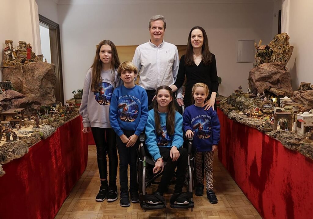 Two adults and four children in Carolina Purpurina sweatshirts pose between the nativity tables; in the centre, Carolina in her wheelchair.