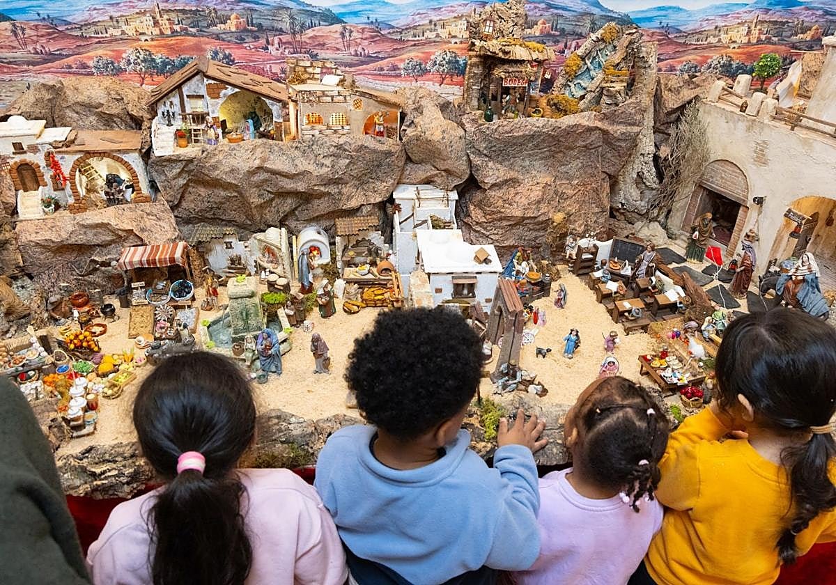 Primeros visitantes del belén: cuatro niños, de espaldas, se asoman a su paisaje de rocas, casas y figuras ante un fondo pintado.