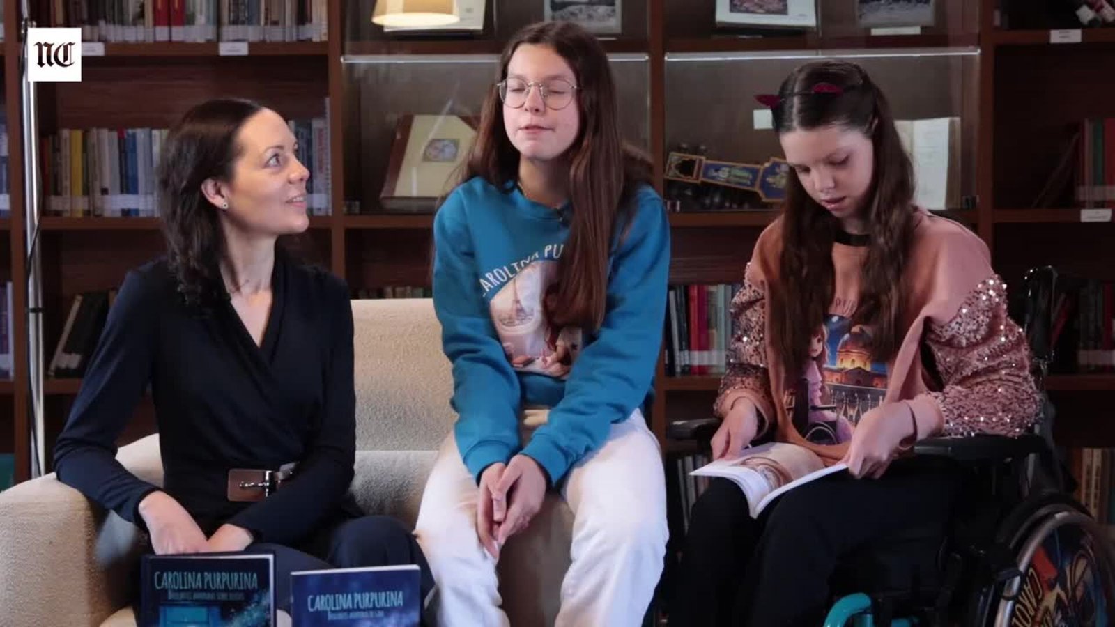 Entrevista por el tercer libro: Laura e Isabel, sentadas ante dos libros de Carolina Purpurina, y Carolina, en su silla de ruedas, hojeando uno.