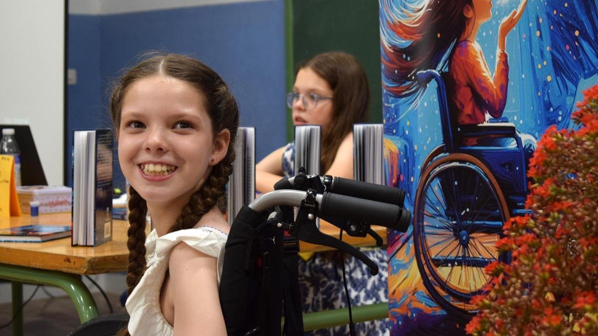 Presentación del libro: Carolina sonríe en su silla de ruedas junto al cartel de la portada; detrás, una niña sentada a la mesa de libros.