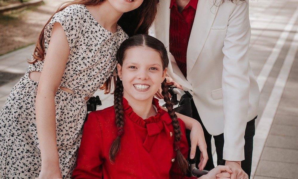 Carolina, con vestido rojo y trenzas, sonríe en su silla de ruedas, abrazada por su familia, cuyos rostros quedan fuera del encuadre.