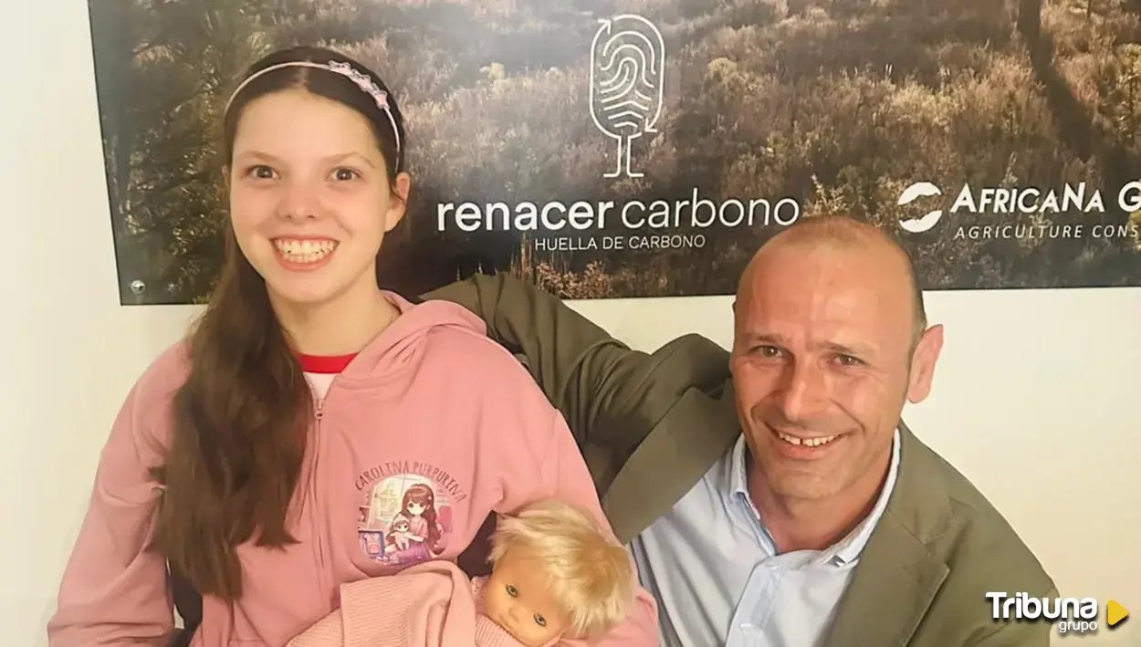 Una niña con sudadera rosa de Carolina Purpurina y un muñeco en brazos posa junto a un hombre ante el cartel de Renacer Carbono.